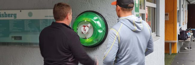 Zwei Personen stehen vor einem Defibrillator an der Wand des Eisplatzes Lindenberg.