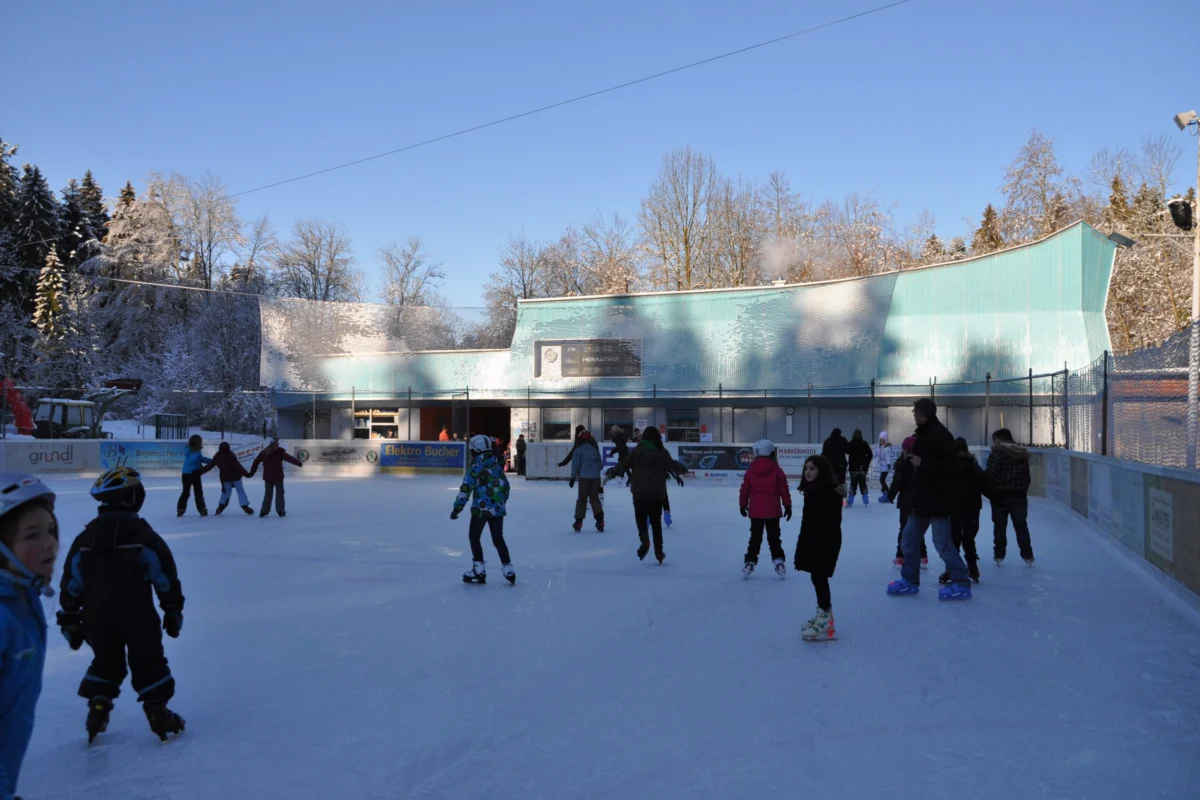 Publikumslauf in Lindenberg Menschen laufen bei sonnigem Wetter auf dem Eisplatz Lindenberg, umgeben von Winterlandschaft und blauen Gebäuden.