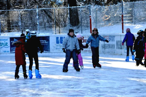 schulsport Kinder fahren Schlittschuh auf der Eisbahn Lindenberg, umgeben von verschneiten Bäumen und Werbebannern.