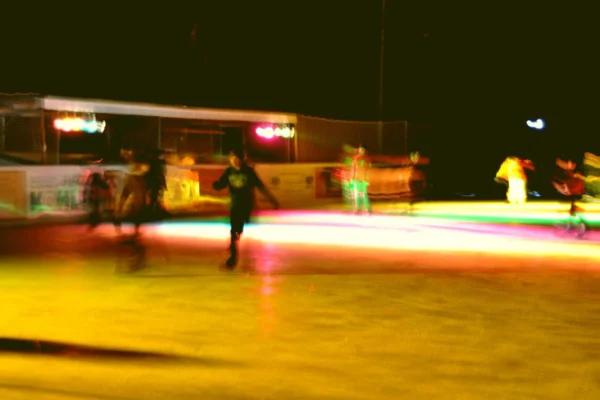 Eisdisco-am-Eisplatz-lindenberg-e1573400605441-everglow Personen beim nächtlichen Eislaufen auf einer farbig beleuchteten Eisfläche, unscharf und lebhaft dargestellt.