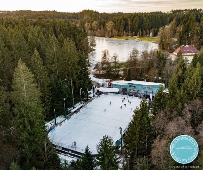 Luftaufnahme des Eisplatzes Lindenberg im Wald, mit Spielern auf der Eisfläche und einem gefrorenen See im Hintergrund.