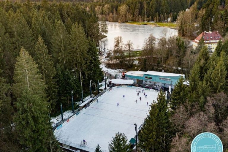 Eisplatz am Waldsee aus der Luft Luftaufnahme des Eisplatzes Lindenberg im Wald, mit Spielern auf der Eisfläche und einem gefrorenen See im Hintergrund.