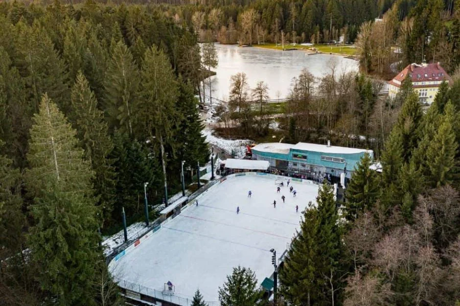 Eisplatz Lindenberg von Oben Luftaufnahme des Eisplatzes Lindenberg, umgeben von Wald, mit Eisläufern auf der Eisfläche und einem nahen See.
