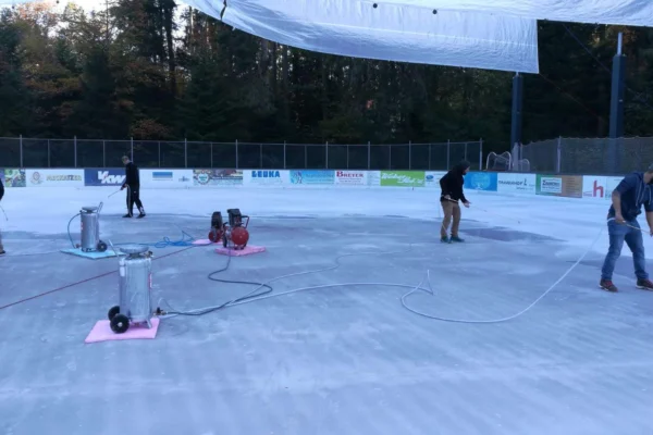 Förderverein bei der Arbeit Personen präparieren mit Schläuchen die Eisfläche am Eisplatz Lindenberg, umgeben von Wald und Werbebannern.