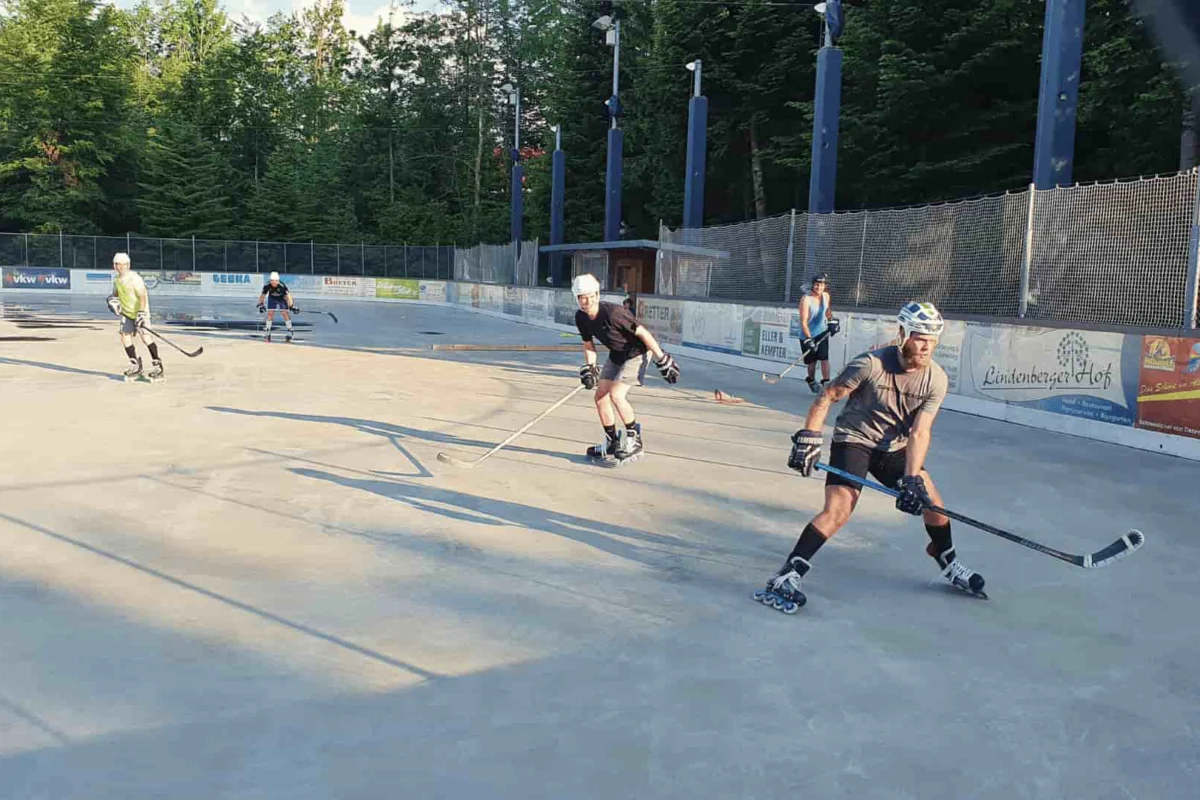 inline-hockey-lindenberg-slider Männer spielen Inline-Hockey bei Sonnenuntergang auf dem Eisplatz Lindenberg, umgeben von Bäumen und Werbebannern.
