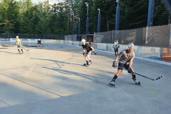 inline-hockey-lindenberg-slider Männer spielen Inline-Hockey bei Sonnenuntergang auf dem Eisplatz Lindenberg, umgeben von Bäumen und Werbebannern.