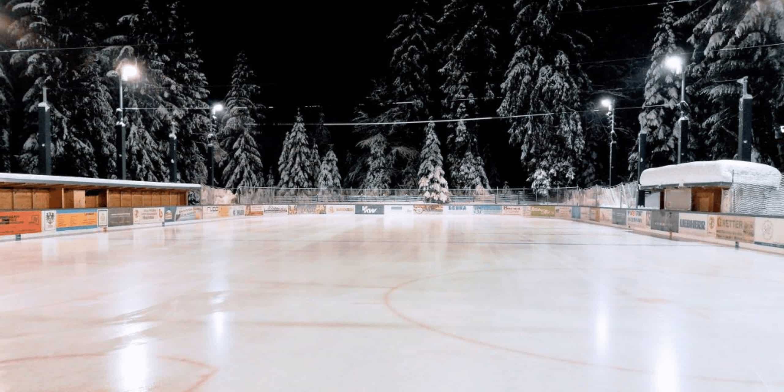 Verschneiter Eisplatz in Lindenberg, umgeben von beleuchteten Bäumen und Holzhütten in winterlicher Nachtstimmung.