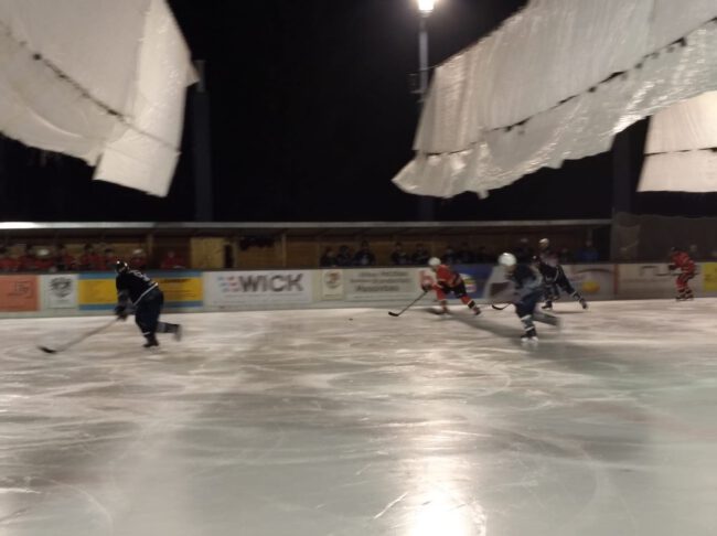Mehrere Eishockeyspieler beim nächtlichen Spiel auf dem Eisplatz Lindenberg, umgeben von Zuschauern.
