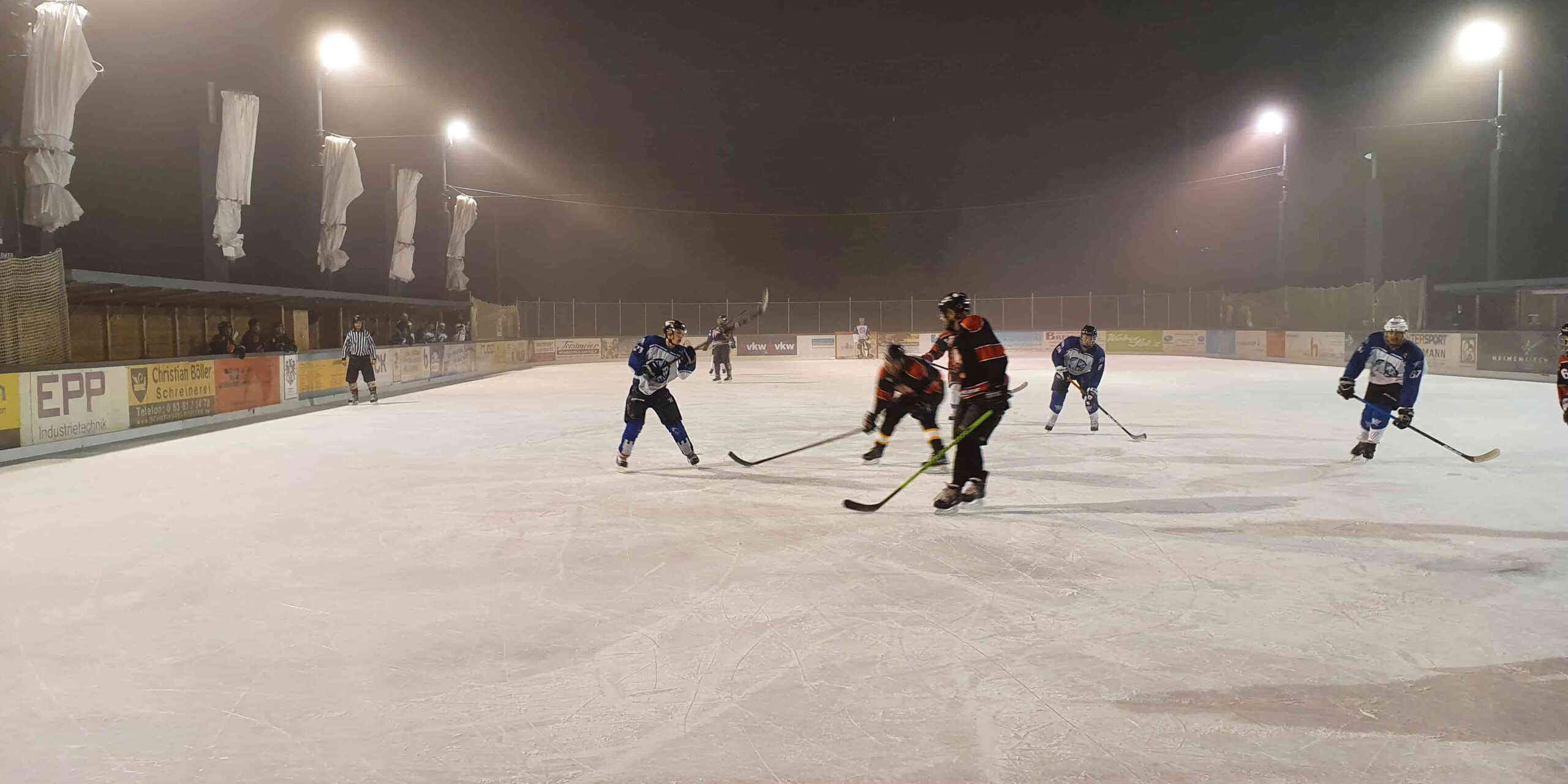 Eishockeyspiel bei Nacht auf dem Eisplatz Lindenberg, Spieler in blauen und roten Trikots, beleuchtete Spielfläche.