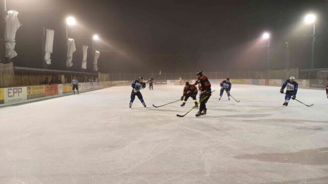 Eishockeyspiel bei Nacht auf dem Eisplatz Lindenberg, Spieler in blauen und roten Trikots, beleuchtete Spielfläche.