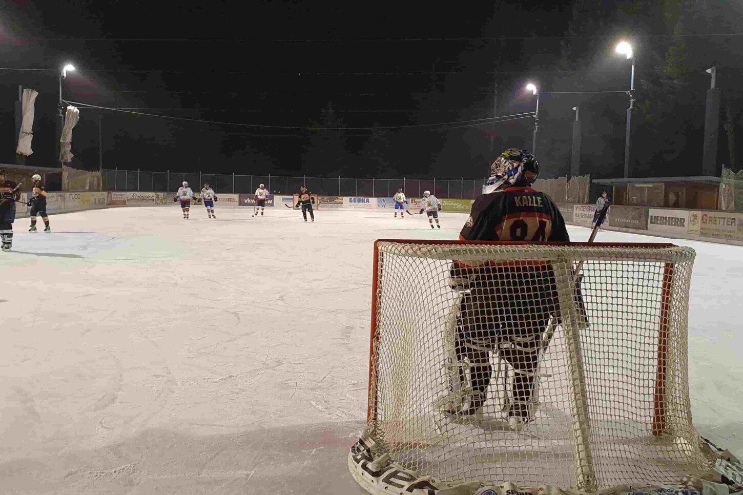 silverlines Nachtspiel auf der Eisbahn Lindenberg, ein Torhüter beobachtet das Eishockeyspiel, Spieler im Hintergrund aktiv.