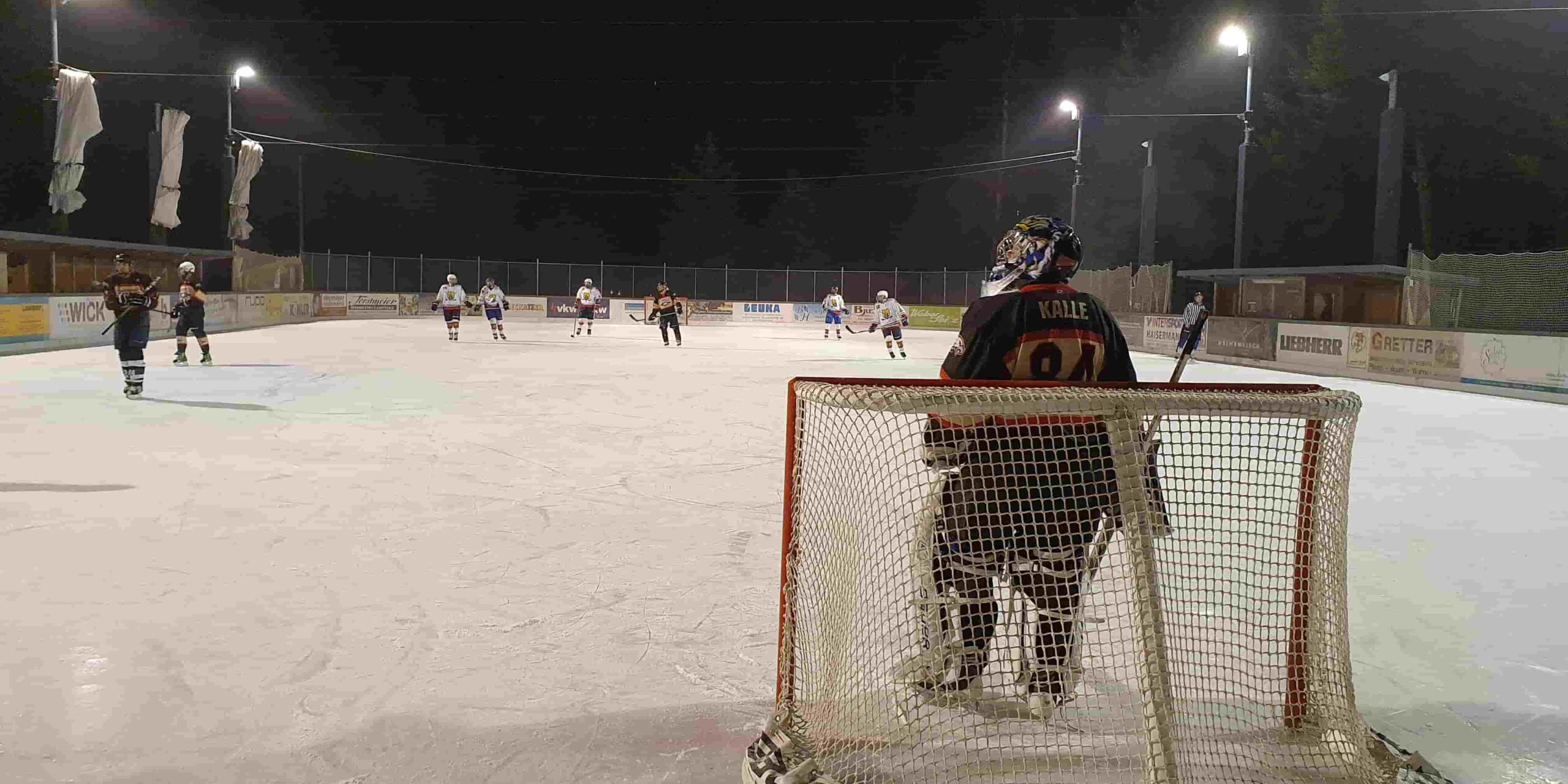 silverlines Nachtspiel auf der Eisbahn Lindenberg, ein Torhüter beobachtet das Eishockeyspiel, Spieler im Hintergrund aktiv.