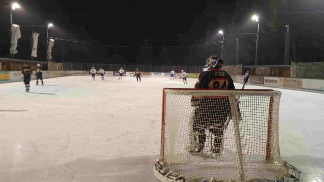 silverlines Nachtspiel auf der Eisbahn Lindenberg, ein Torhüter beobachtet das Eishockeyspiel, Spieler im Hintergrund aktiv.