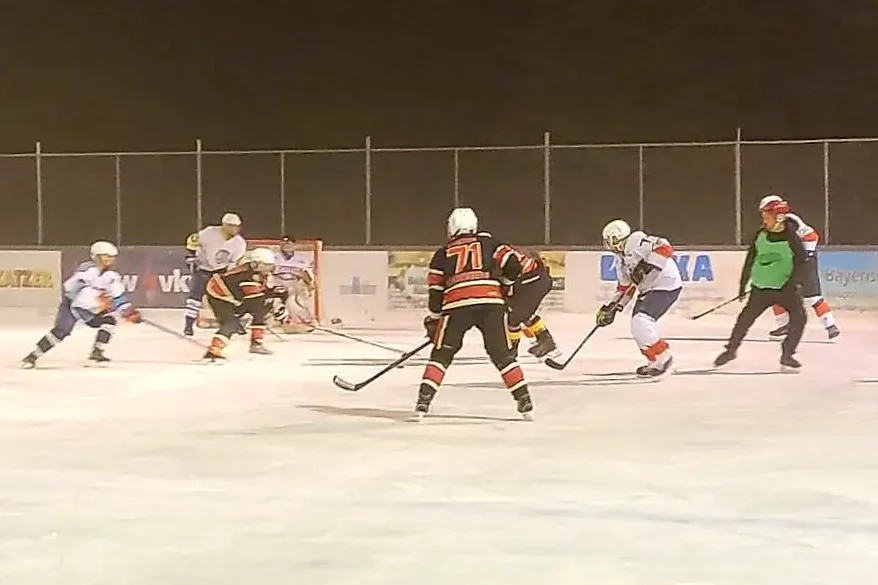 wp-16376066910971944333033173623629 Eishockeyspieler im Wettkampf auf dem Eisplatz Lindenberg bei Nacht, umgeben von Werbebannern, intensives Spielgeschehen.