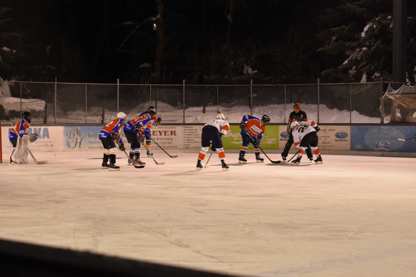 wangen-weiherwings-2021 Eishockeyspiel auf beleuchtetem Eisplatz mit Spielern in blauen und weißen Trikots, unter den Augen eines Schiedsrichters.