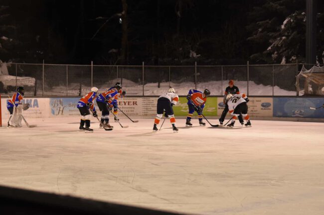 wangen-weiherwings-2021 Eishockeyspiel auf beleuchtetem Eisplatz mit Spielern in blauen und weißen Trikots, unter den Augen eines Schiedsrichters.