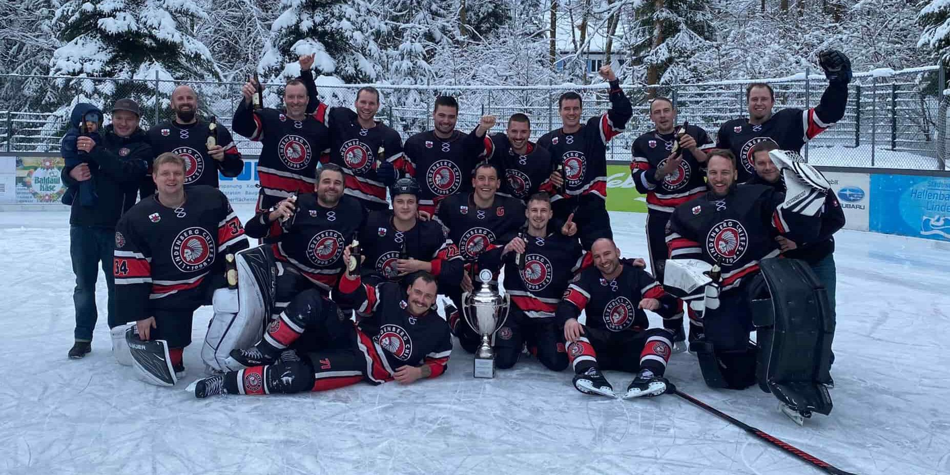 Eine Eishockeymannschaft feiert auf dem Eis mit einem großen Pokal, umgeben von verschneiten Bäumen.
