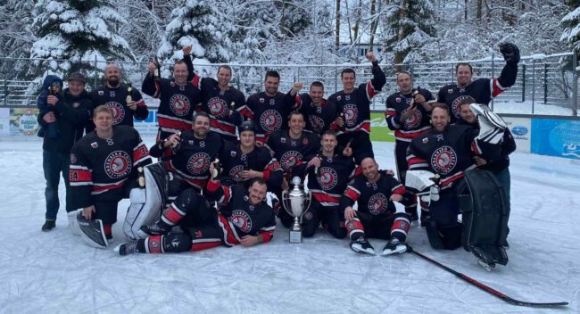 Eine Eishockeymannschaft feiert auf dem Eis mit einem großen Pokal, umgeben von verschneiten Bäumen.