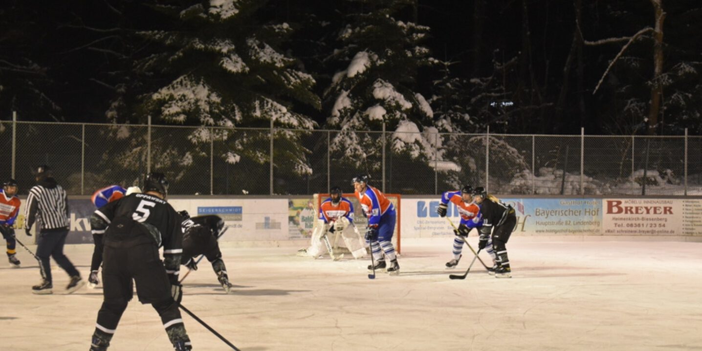 Eishockeyspiel auf dem Eisplatz Lindenberg, Teams in blauer und schwarzer Ausrüstung vor schneebedeckten Tannen.