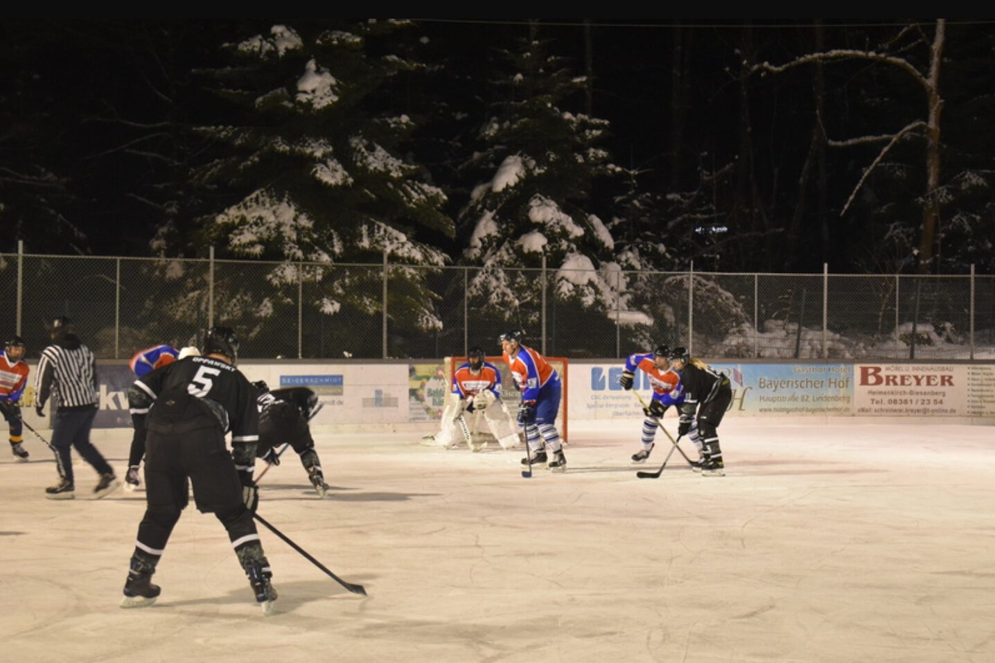 image_editor_output_image-1281726452-1642546978406.jpg Eishockeyspiel auf dem Eisplatz Lindenberg, Teams in blauer und schwarzer Ausrüstung vor schneebedeckten Tannen.