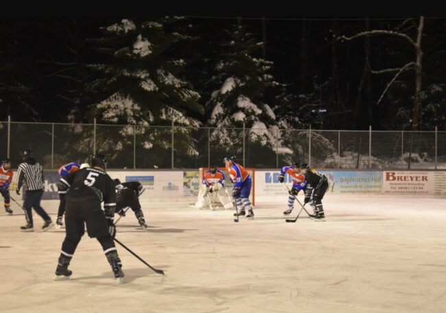 Eishockeyspiel auf dem Eisplatz Lindenberg, Teams in blauer und schwarzer Ausrüstung vor schneebedeckten Tannen.