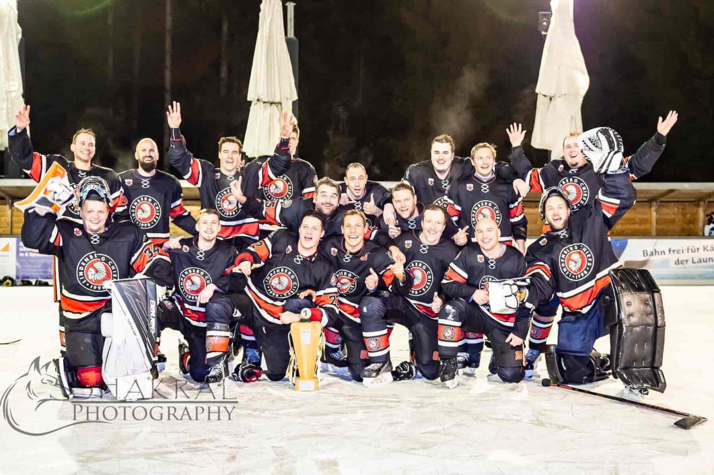 chiefs-meister-2022 Auf dem Eis feiern die Chiefs 2022 Meisterschaftssieg, das Team posiert jubelnd in schwarzen Trikots mit Pokal.
