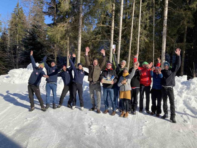 Eine Gruppe fröhlicher Menschen freut sich im Schnee, wohl nach einem Eisstockturnier in Lindenberg. Bäume im Hintergrund.