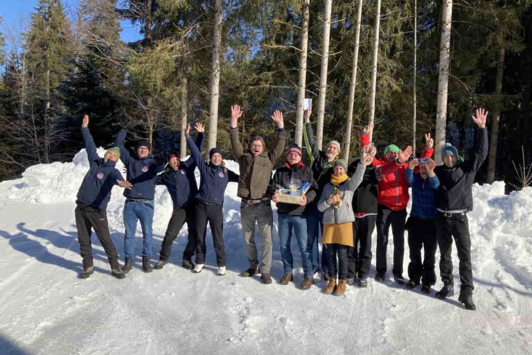 eisstockturnier-eisplatz-lindenberg-2022 Eine Gruppe fröhlicher Menschen freut sich im Schnee, wohl nach einem Eisstockturnier in Lindenberg. Bäume im Hintergrund.