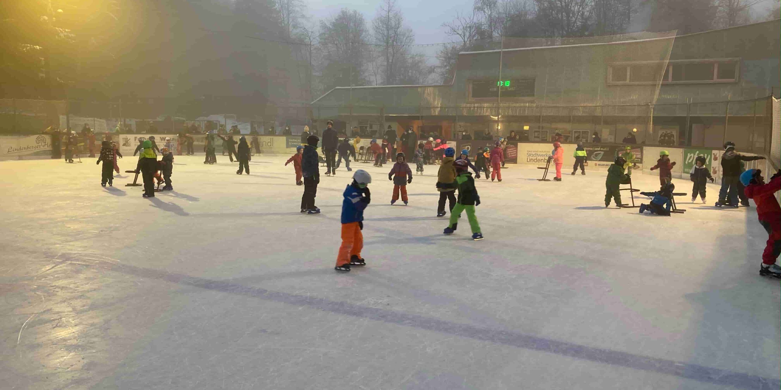 Publikumslauf Kinder und Erwachsene eislaufen auf dem Eisplatz Lindenberg an einem nebligen Tag, umgeben von Bäumen und Gebäuden.