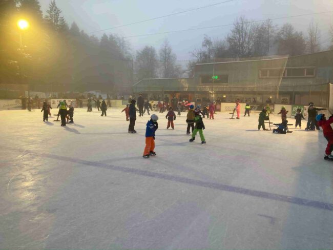 Kinder und Erwachsene eislaufen auf dem Eisplatz Lindenberg an einem nebligen Tag, umgeben von Bäumen und Gebäuden.