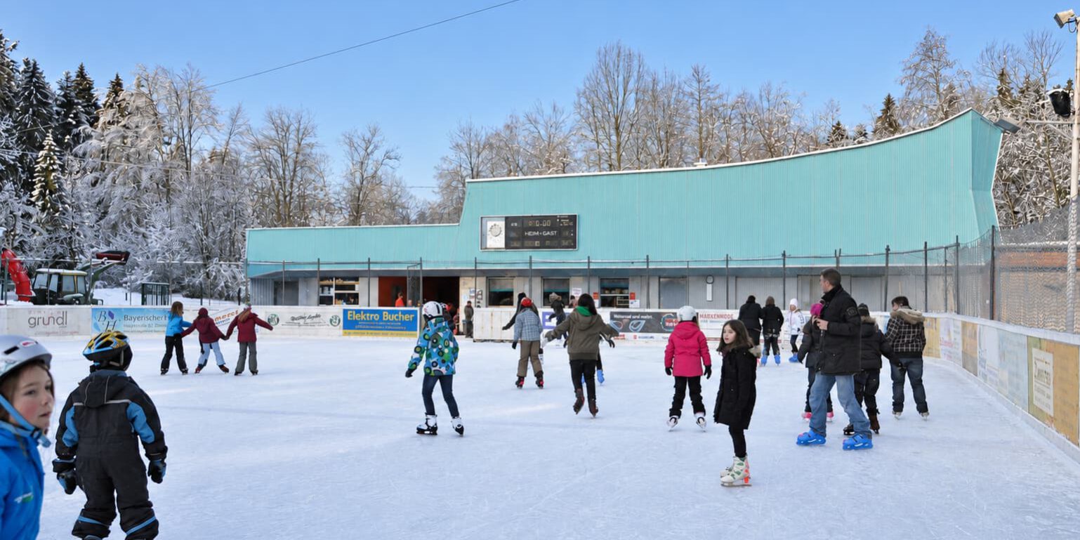 Kinder lernen im Kunsteisstadion Lindenberg Schlittschuhlaufen, umgeben von weiteren Eisläufern.