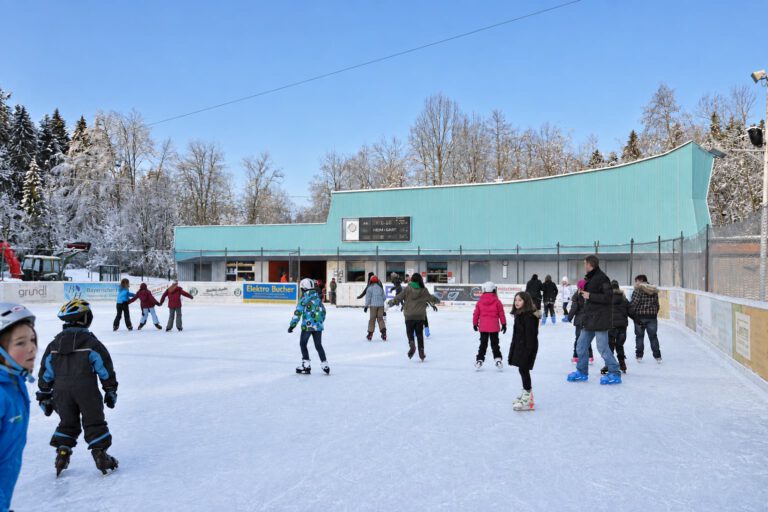 Kinder lernen im Kunsteisstadion Lindenberg Schlittschuhlaufen, umgeben von weiteren Eisläufern.