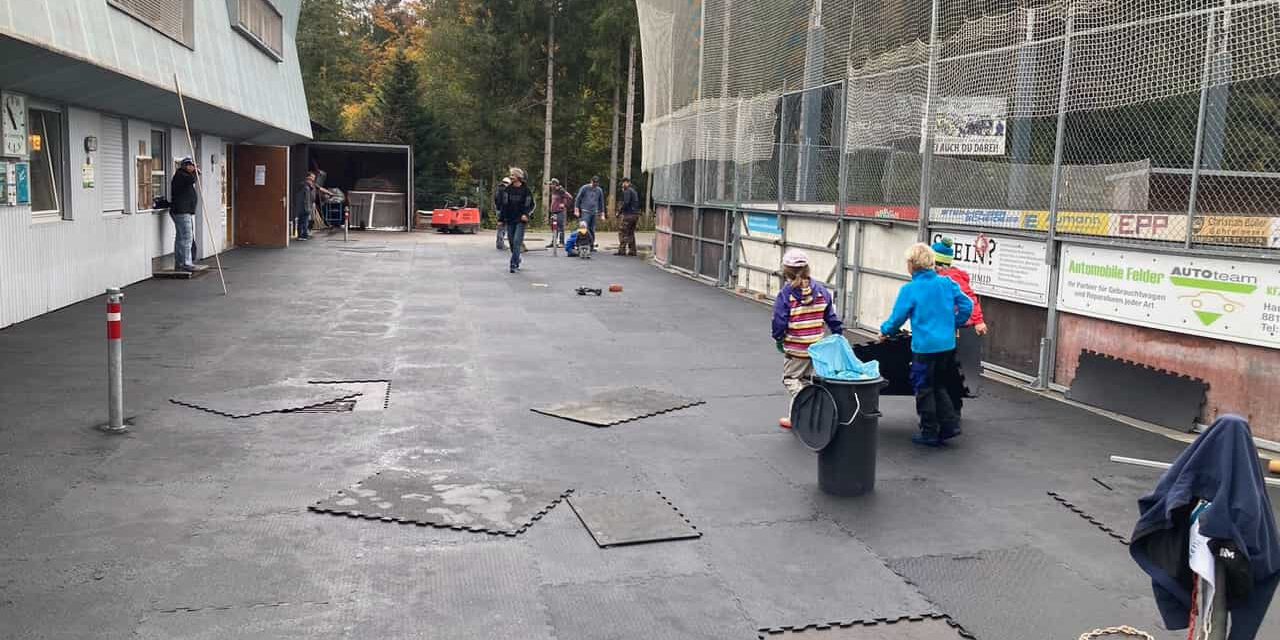 eisplatz-mattenlegen-förderverein Menschen legen Matten auf dem Eisplatz Lindenberg aus; Kinder helfen eifrig mit, während Erwachsene zuschauen.