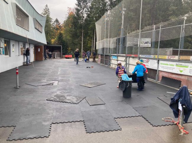 Menschen legen Matten auf dem Eisplatz Lindenberg aus; Kinder helfen eifrig mit, während Erwachsene zuschauen.