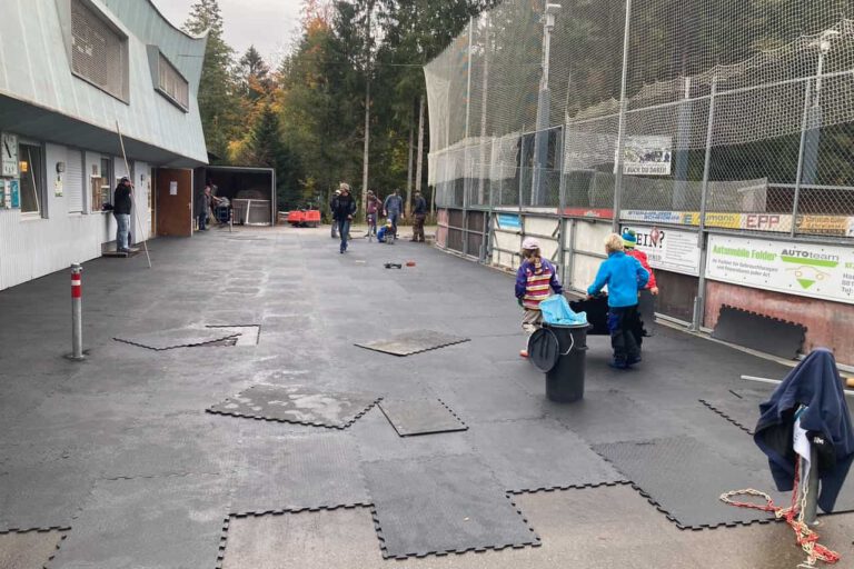 eisplatz-mattenlegen-förderverein Menschen legen Matten auf dem Eisplatz Lindenberg aus; Kinder helfen eifrig mit, während Erwachsene zuschauen.