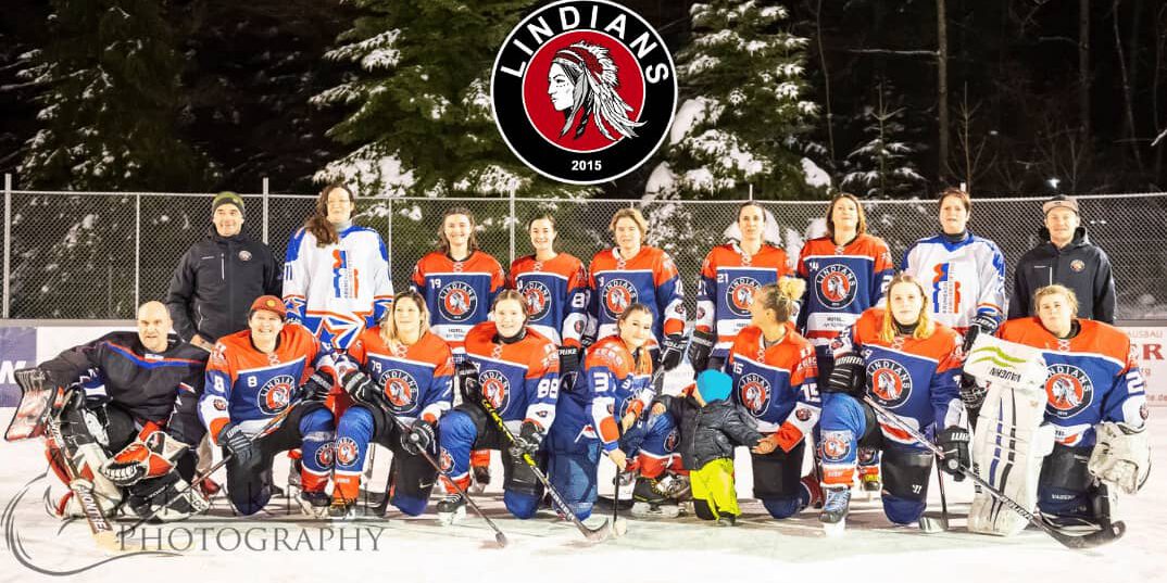 Eishockey-Team Lindians posiert in orangen Trikots auf Eisfläche unter dem Logo, umgeben von schneebedeckten Bäumen.