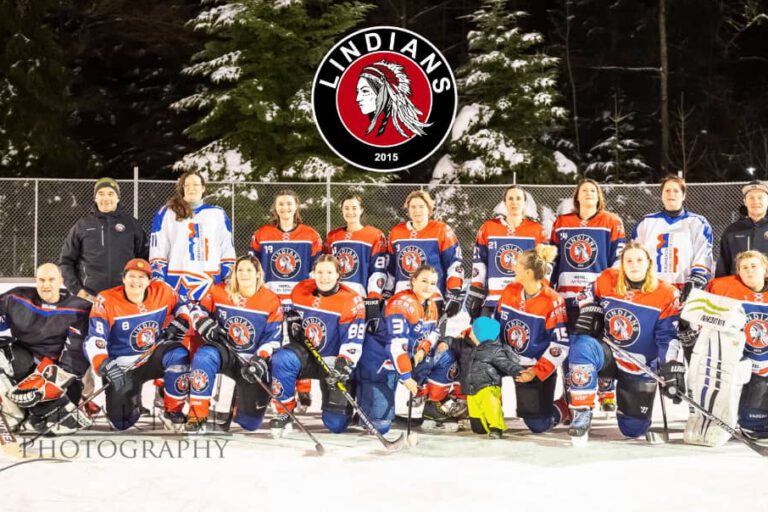 Lindians Damen-Eishockeymannschaft Eishockey-Team Lindians posiert in orangen Trikots auf Eisfläche unter dem Logo, umgeben von schneebedeckten Bäumen.