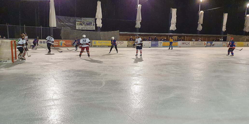 Eishockeyspiel unter Flutlicht auf der Eisfläche in Lindenberg, Spieler in Aktion mit Zuschauern im Hintergrund.