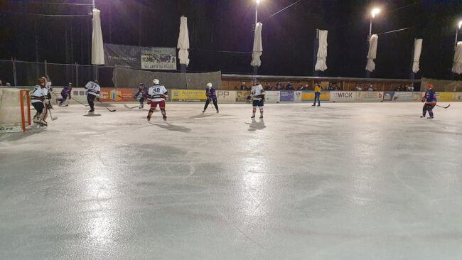 Eishockeyspiel unter Flutlicht auf der Eisfläche in Lindenberg, Spieler in Aktion mit Zuschauern im Hintergrund.