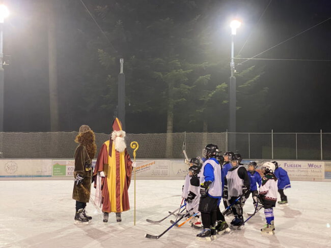 nikolaus am Eisplatz Lindenberg Nikolaus und Knecht Ruprecht begrüßen eine Gruppe von Kindern im Eishockey-Outfit auf einer mit Flutlicht beleuchteten Eisbahn.