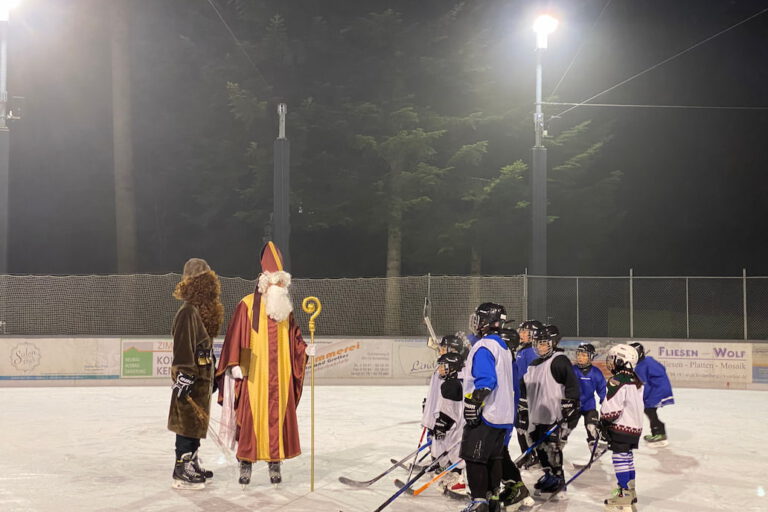 nikolaus am Eisplatz Lindenberg Nikolaus und Knecht Ruprecht begrüßen eine Gruppe von Kindern im Eishockey-Outfit auf einer mit Flutlicht beleuchteten Eisbahn.