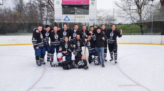 Eishockeyteam Black Flyers auf dem Eisplatz Lindenberg, fröhlich posierend vor der Anzeigetafel im Freien.