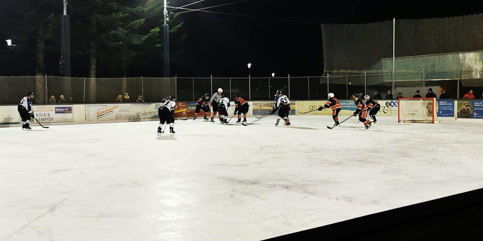 Eishockey-Spiel bei Nacht auf dem Eisplatz Lindenberg, Spieler kämpfen um den Puck, Zuschauer am Spielfeldrand.