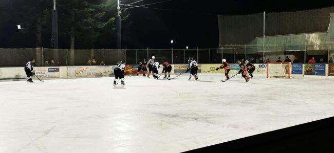 Eishockey-Spiel bei Nacht auf dem Eisplatz Lindenberg, Spieler kämpfen um den Puck, Zuschauer am Spielfeldrand.
