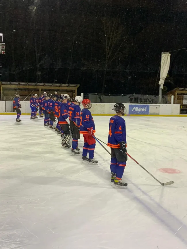 Eishockeyspieler in blauen Trikots stehen in einer Reihe auf dem Eisplatz Lindenberg unter dunklem Himmel.