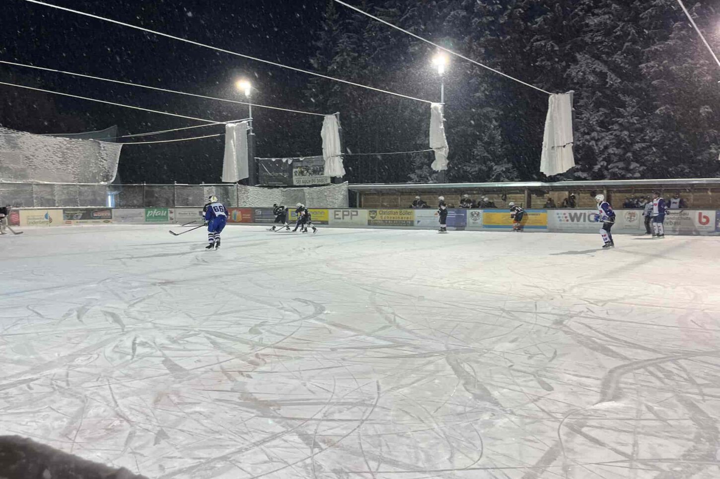 mtu-friedrichshafen-black-flyers-ravensburg-wael-2023 Eishockeyspiel auf dem Eisplatz Lindenberg bei Nacht, Spieler in blauen und schwarzen Trikots auf verschneitem Feld.
