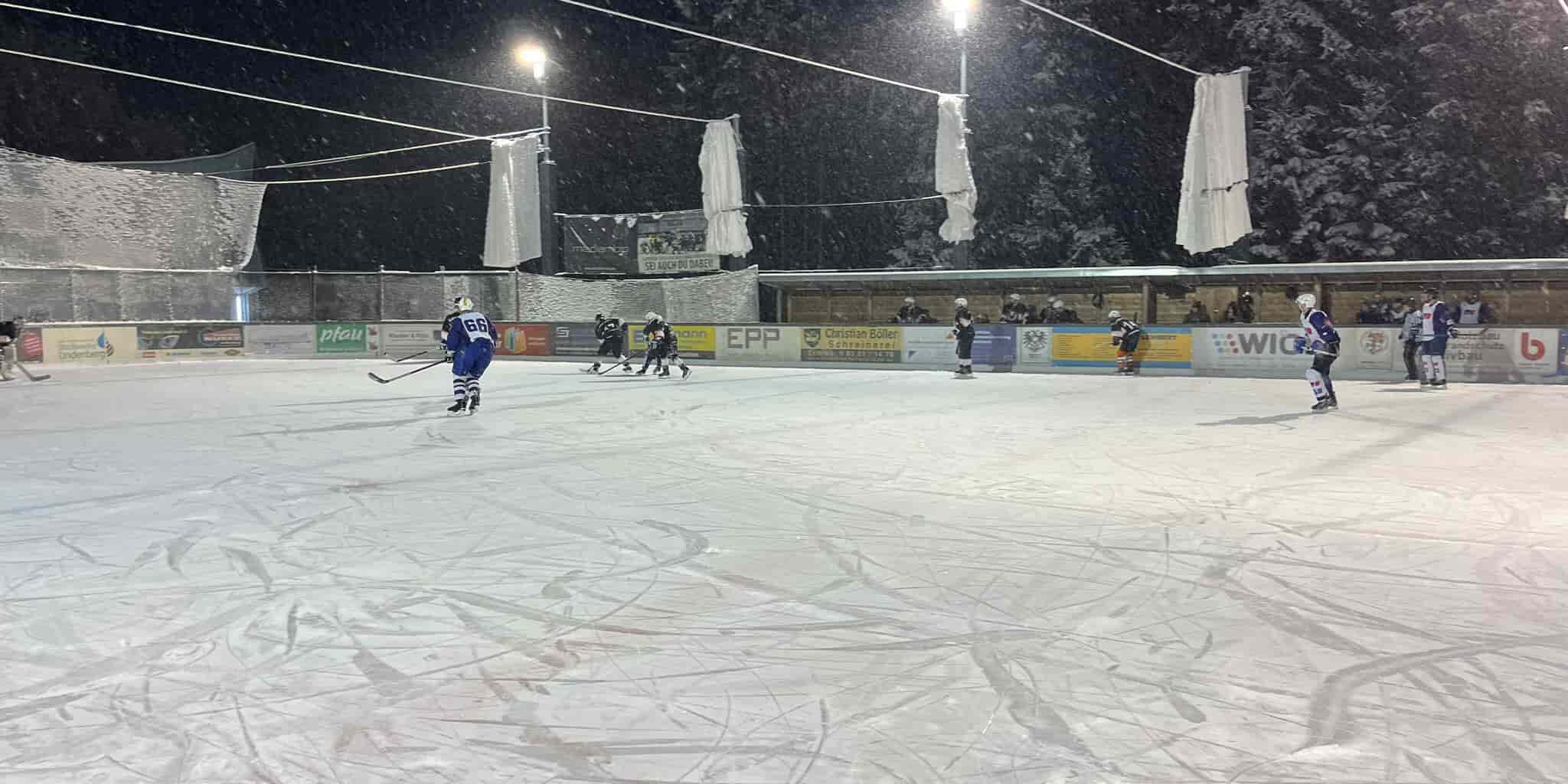 mtu-friedrichshafen-black-flyers-ravensburg-wael-2023 Eishockeyspiel auf dem Eisplatz Lindenberg bei Nacht, Spieler in blauen und schwarzen Trikots auf verschneitem Feld.
