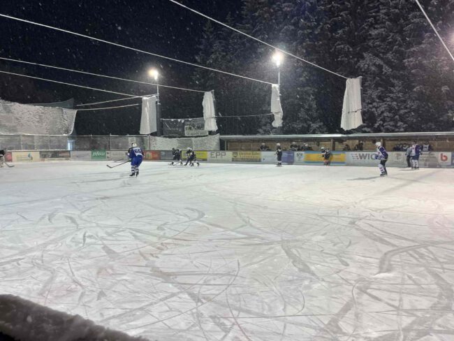mtu-friedrichshafen-black-flyers-ravensburg-wael-2023 Eishockeyspiel auf dem Eisplatz Lindenberg bei Nacht, Spieler in blauen und schwarzen Trikots auf verschneitem Feld.
