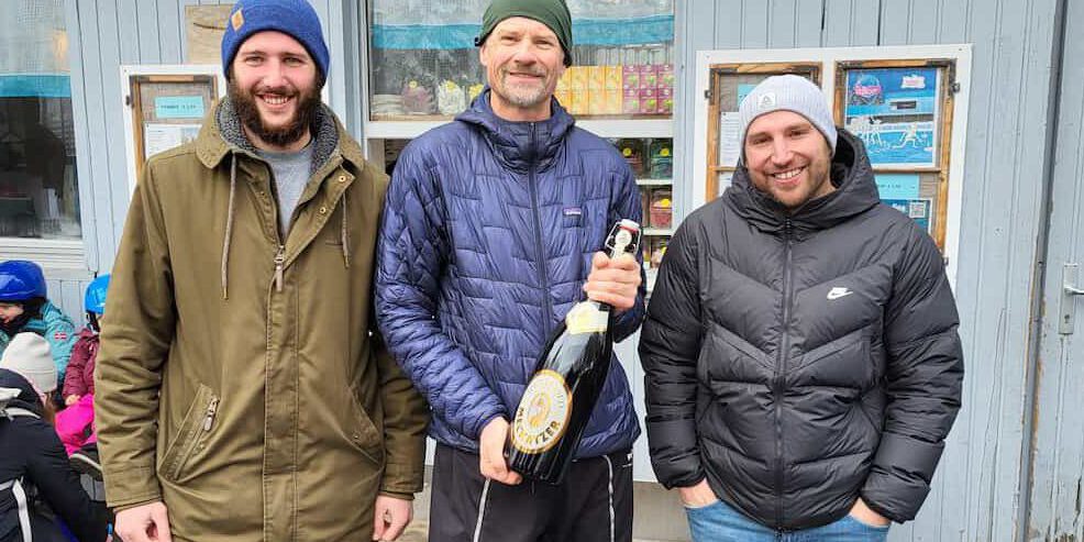 Drei Männer posieren vor einem Gebäude, der mittlere hält eine große Flasche mit Aufschrift "Walter".