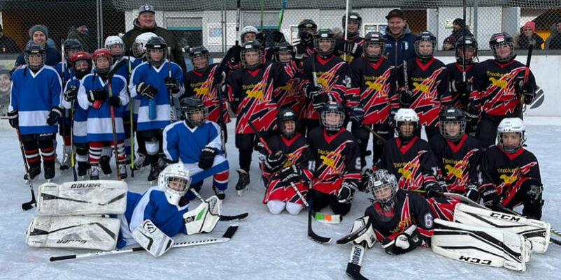Kinder-Eishockeyteams in blauen und schwarzen Trikots posieren gemeinsam auf dem Eisplatz von Lindenberg.
